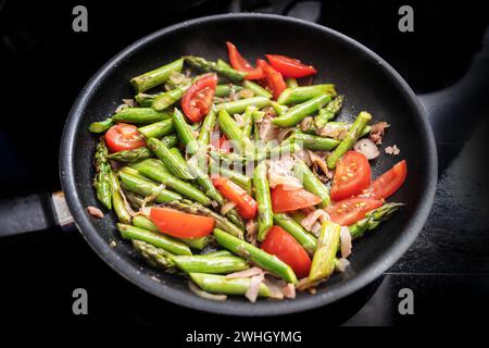 Grüner Spargel, Tomaten und Parmaschinken in einer schwarzen Bratpfanne auf dem Herd, die eine gesunde Frühjahrsmahlzeit kocht, ausgewählte Focu Stockfoto