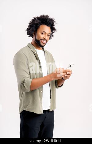 Lächelnder afrikanischer Mann aus den 30er Jahren in Freizeitkleidung mit Smartphone isoliert auf weiß stehend, glücklicher Kerl online SMS, Websurfen, männlicher Student genießt Online-Chats, mit mobiler App Stockfoto