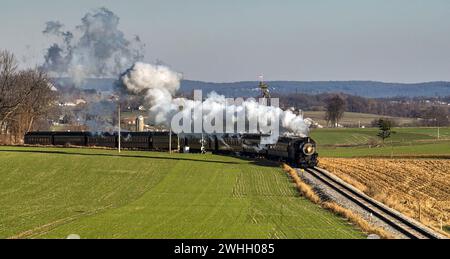 Drohnenansicht eines antiken Dampfeisenzugs, der sich durch die Landschaft und das Ackerland nähert und Rauch bläst Stockfoto