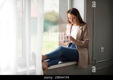 Work-Life-Balance, kaukasische weibliche Managerin auf der Fensterbank im Flur mit Smartphone Stockfoto
