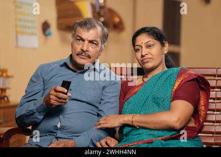 Indische ältere ältere Ehepaare schauen fernsehen oder Fernsehen zu Hause mit Fernbedienung - Konzept des Ruhestandslebens, Beziehungsbindung und Stockfoto