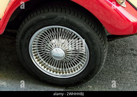 Das Bild zeigt eine Nahaufnahme eines Fahrzeugrads mit einer glänzenden, komplizierten Radkappe mit Drahtspeichen. Das Auto hat eine rote Karosserie und steht auf Asphalt. Stockfoto