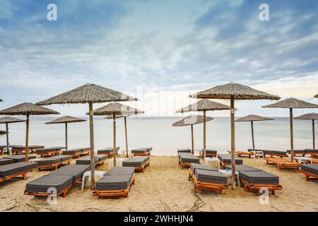 Hölzerne Strandliegen mit weichen Kissen und Strohschirmen direkt am Meer in einem Touristenort in Griechenland vor bewölktem Wetter Stockfoto