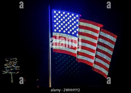 Nächtliche Darstellung von drei amerikanischen Flaggen aus hellen Lichtern, die auf Einem Stab montiert sind, mit Einer Weihnachtsbaumbeleuchtung, die auf der linken Seite sichtbar ist. Stockfoto