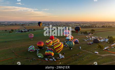 Eine Luftaufnahme von einer Gruppe von bunten Heißluftballons, die sich auf den Flug in der Dämmerung vorbereiten, mit Zuschauern und Fahrzeugen, die sich auf Einem ländlichen Feld versammelt haben. Stockfoto