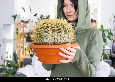 Großer echinocactus Gruzoni in den Händen einer Frau im Inneren eines Gewächshauses mit Regalsammlungen von Hauspflanzen Stockfoto