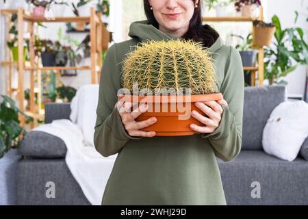 Großer echinocactus Gruzoni in den Händen einer Frau im Inneren eines Gewächshauses mit Regalsammlungen von Hauspflanzen Stockfoto