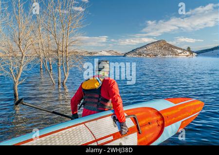 Ein männlicher Paddler, der einen Trockenanzug, eine Schwimmweste und eine Sicherheitsleash trägt, beginnt mit dem Training auf einem langen Rennstativ am Paddleboard auf einem Bergsee in Colorado Stockfoto