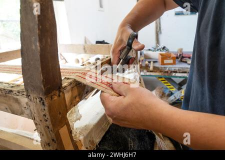 Polsterer mit traditioneller Handwerkskunst verkürzt einen neuen Riemen an einem alten Stuhl mit Schere in seiner Werkstatt, Kopierraum, auswählen Stockfoto