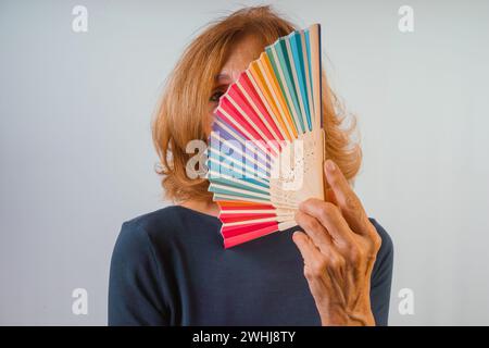 Senior Fit kaukasische 60er 70er Jahre selbstbewusste Frau, die Regenbogenfan über weißem Hintergrund hält und ein blaues Langarmhemd trägt Stockfoto