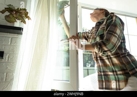 Die Frau wäscht das Fenster des Hauses manuell mit einem Lappen mit Sprühreiniger und wischt das Innere mit weißen Vorhängen. Res Stockfoto