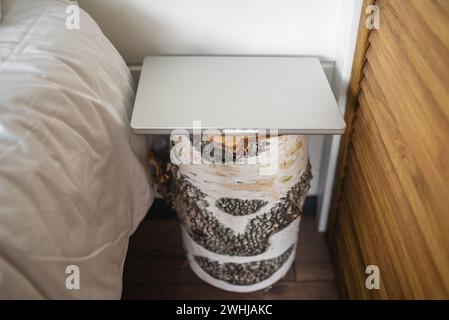 Ein einzigartiger Nachttisch aus Birkenholz steht neben einem Bett mit geschlossenem Laptop auf der Oberseite und verbindet rustikalen Charme mit modernem Komfort in einem gemütlichen Schlafzimmer. Der Laptop liegt auf dem Nachttisch neben dem Bett Stockfoto