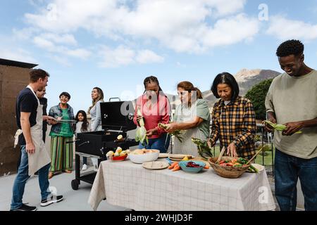 Generationenübergreifende Freunde, die Spaß beim Grillen auf dem Dach des Hauses haben - fröhliche, multirassische Menschen, die zusammen kochen Stockfoto