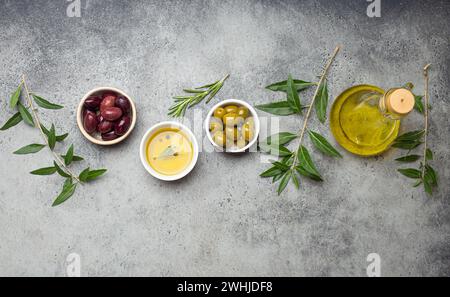 Zusammensetzung mit grünen und braunen Oliven, nativem Olivenöl extra in Glasflasche, Olivenbaumzweige auf grauem Betonsteinrost Stockfoto