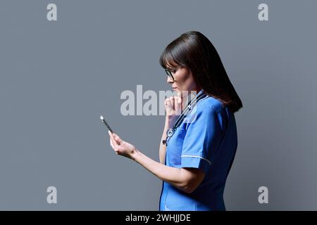 Krankenschwester mit Smartphone, Profilansicht auf grauem Atelierhintergrund Stockfoto