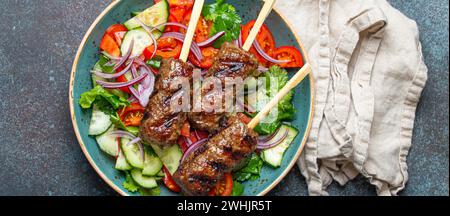 Gegrillte Fleischspieße Rindfleisch-Kebabs auf Stäbchen serviert mit frischem Gemüsesalat auf Teller auf rustikalem Betonhintergrund von oben. Stockfoto