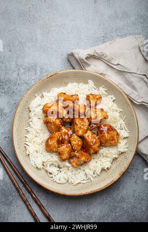 Chinesisches süß-saures, klebriges Huhn mit Sesamsamen und Reis auf Keramikplatte mit Essstäbchen von oben, graues rustikales Steinbaa Stockfoto