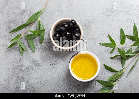 Schwarze Oliven und extra natives Olivenöl in einer kleinen Schüssel mit Olivenbaumzweigen von oben auf grauem Betonhintergrund Stockfoto
