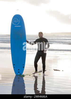 Surfer steht mit seinem Surf Board im Meer bei Sonnenuntergang mit einer Insel dahinter in Essaouira, Marokko. Februar 2024 Stockfoto