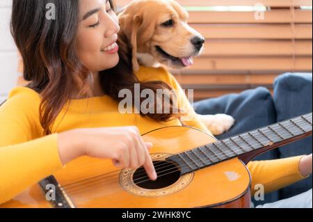 An einem Wochenende spielen Eine junge Asiatin und ihr Beagle-Welpe Gitarre und singen fröhlich zusammen im Wohnzimmer Stockfoto