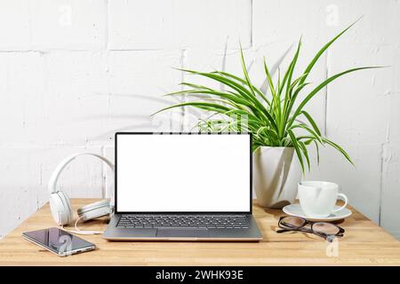 Holzschreibtisch mit offenem Laptop-Modell, Kopfhörer, Telefon, Brille und Pflanze vor einer weiß gestrichenen Wand, Büro, Büro, Büro Stockfoto