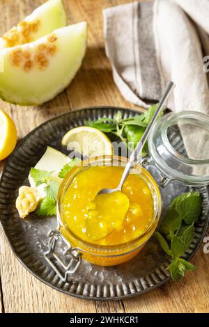 Hausgemachtes Preserve. Süße Melone und Zitrusmarmelade oder Gelee in einem kleinen Glasgefäß mit frischen Melonenscheiben auf einem rustikalen Holztisch. Stockfoto