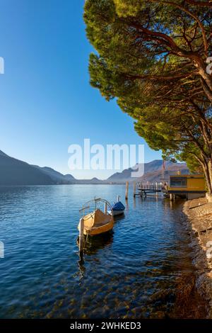 Wunderschöner Jetty und altes Fischerboot mit Bäumen an der Uferpromenade am Luganersee an einem sonnigen Tag mit Berg in Morcote, Tessin, Schweiz Stockfoto