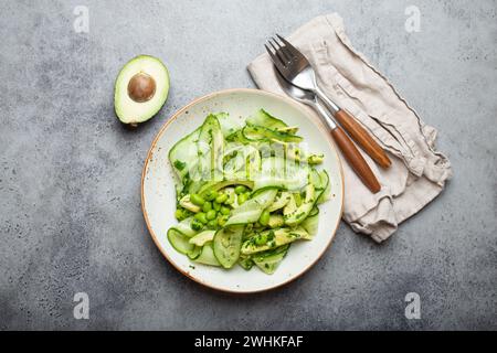 Gesunde vegane grüne Avocado Salatschüssel mit geschnittenen Gurken, Edamame Bohnen, Olivenöl und Kräutern auf Keramikplatte Draufsicht, grau Stockfoto