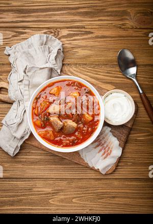 Ukrainischer Borschtsch, rote Bete Suppe mit Fleisch, in weißer Schüssel mit Sauerrahm und salobscheiben, rustikaler Holzuntergrund. Traditi Stockfoto