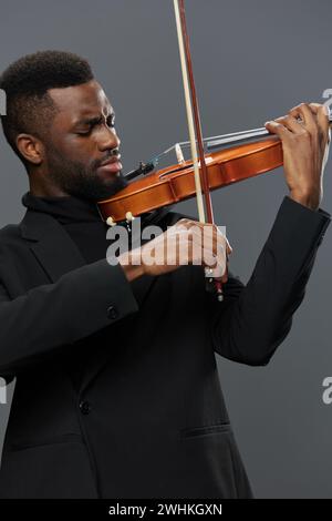 Gefühlvoller afroamerikanischer Mann in schwarzem Anzug, der Geige auf grauem Hintergrund in eleganter musikalischer Performance spielt Stockfoto