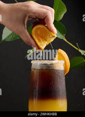 Eiskaffee mit Orangensaft in einem durchsichtigen Glas, die Hand einer Frau drückt eine Orangenscheibe in ein Seifenblasengetränk Stockfoto