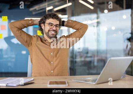 Gut gemacht Geschäftsmann hat das Arbeitsprojekt im Büro am Arbeitsplatz erfolgreich abgeschlossen, Mann mit Händen hinter dem Kopf, der glücklich lächelnd auf den Laptop-Bildschirm blickt, zufrieden mit den Ergebnissen Stockfoto