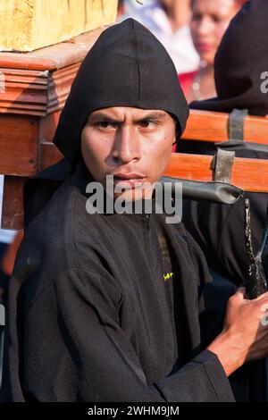 Antigua, Guatemala. Ein schwarzer Cucurucho begleitet einen Karfreitagswagen in einer religiösen Prozession während der Karwoche, La Semana Santa. Stockfoto