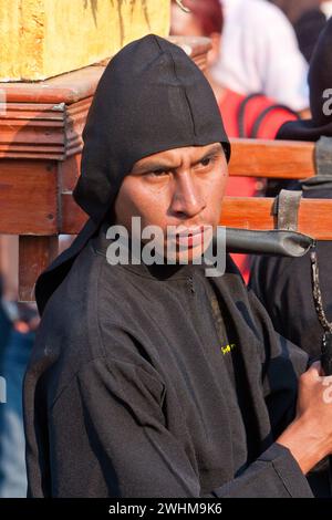 Antigua, Guatemala. Ein schwarzer Cucurucho begleitet einen Karfreitagswagen in einer religiösen Prozession während der Karwoche, La Semana Santa. Stockfoto