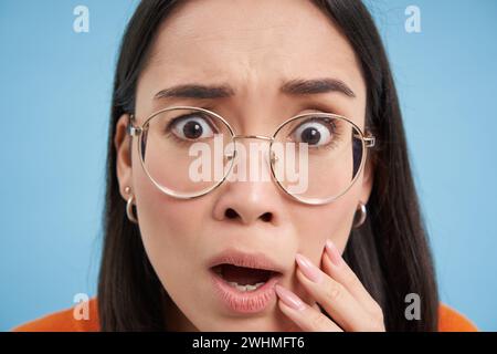 Nahaufnahme eines japanischen Mädchens mit Brille, schockiert und besorgt vor der Kamera, erschreckt, besorgt, steht über blauem Hintergrund Stockfoto