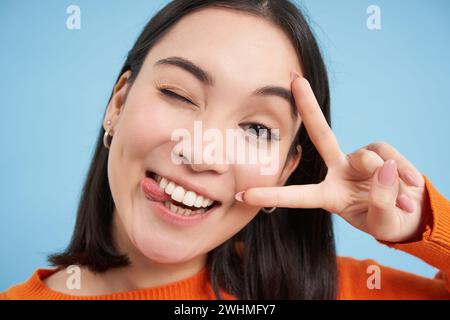 Nahaufnahme des Porträts einer positiven asiatischen Frau, zwinkert und zeigt Peave, V-Zeichen-Geste, klebt Zunge und Lächeln, steht über blauem Bac Stockfoto