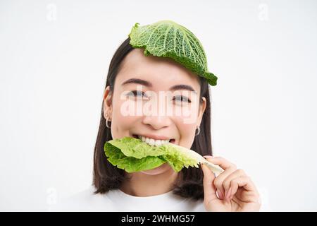 Porträt eines asiatischen Mädchens mit Blättern auf dem Kopf, isst Kohl und lächelt, isoliert auf weißem Hintergrund Stockfoto