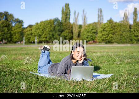 Porträt eines jungen, glücklichen asiatischen Mädchens, das im Park auf einer Decke liegt, Videos ansieht und auf ihrem Laptop im Internet surft Stockfoto