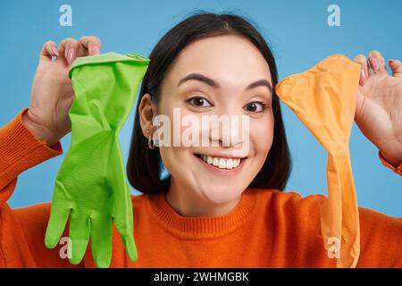 Nahaufnahme Porträt einer lächelnden asiatischen Frau, die zwei verschiedene Latexhandschuhe zum Putzen zeigt, enthusiastisch aussieht, Hausfehler macht Stockfoto