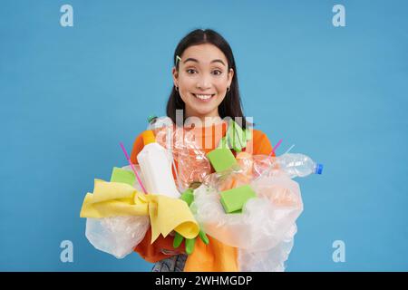 Lächelnde koreanische Frau, recycelt, hält Plastikabfälle und sortiert sie für ein Recycling-Center, blauer Hintergrund Stockfoto