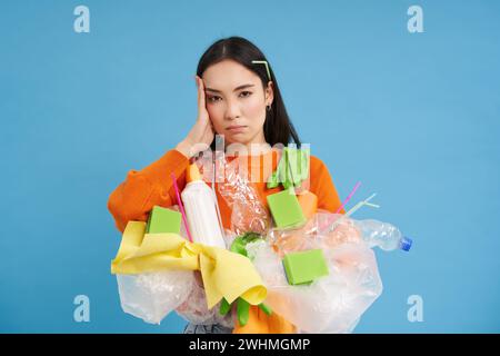 Verärgerte junge Frau, die das Haus putzt, Plastikmüll für das Recycling hält, müde vom Sammeln von Abfall, steht über blauem Hintergras Stockfoto