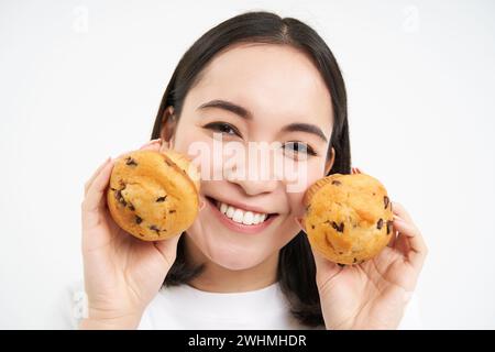 Bild einer koreanischen Frau, die gerne Gebäck isst, mit zwei leckeren Cupcakes im Gesicht und einem Lächeln auf weißem Hintergrund Stockfoto