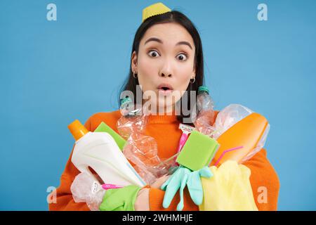 Nahaufnahme eines Mädchens mit überraschtem Gesicht, hält leere Plastikflaschen und grünen Abfall, sammelt Müll zum Recycling, blauer Hinterkopf Stockfoto