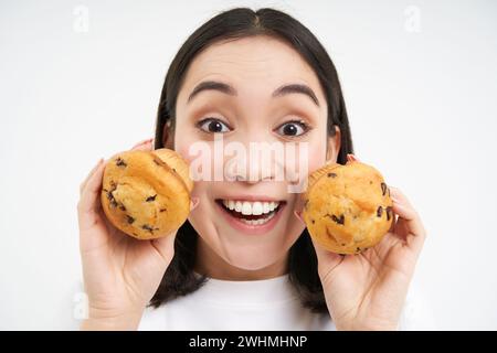 Nahaufnahme Porträt einer asiatischen Frau, zeigt zwei Cupcakes im Gesicht und lächelt, mag Gebäck, isst gerne Bäckerei, weißer Hintergrund Stockfoto