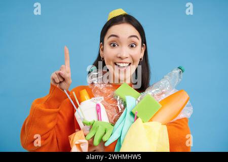 Glückliche junge Frau, die nach oben zeigt, leere Plastikflaschen und Müll zum Recycling hält, Öko-Banner zeigt, Sortierstation adve Stockfoto