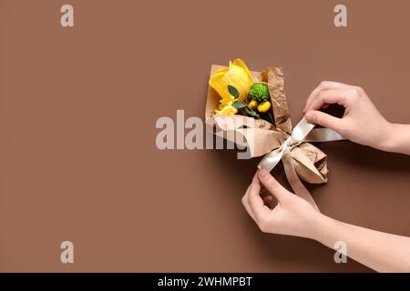 Weibliche Hände mit Ministrauß von schönen gelben Frühlingsblumen im Geschenkpapier auf braunem Hintergrund. Internationaler Frauentag Stockfoto