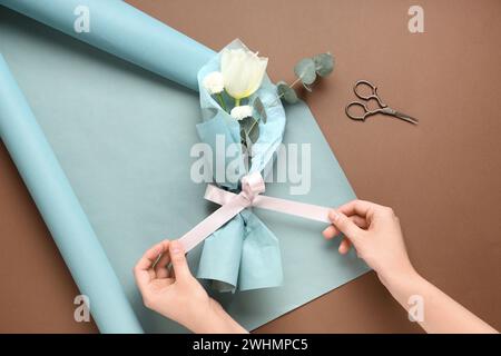 Weibliche Hände mit Ministrauß von schönen Frühlingsblumen in Geschenkpapier und Schere auf braunem Hintergrund. Internationaler Frauentag Stockfoto