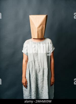 Frau im Kleid mit einer Papiertüte auf dem Kopf, die über dunklem Hintergrund steht Stockfoto