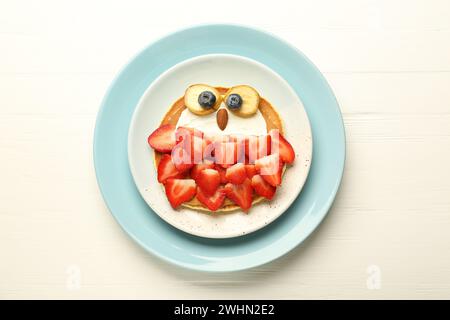 Kreatives Servieren für Kinder. Teller mit süßer Eule aus Pfannkuchen, Erdbeeren, Creme, Banane und Mandel auf weißem Holztisch, Blick von oben Stockfoto