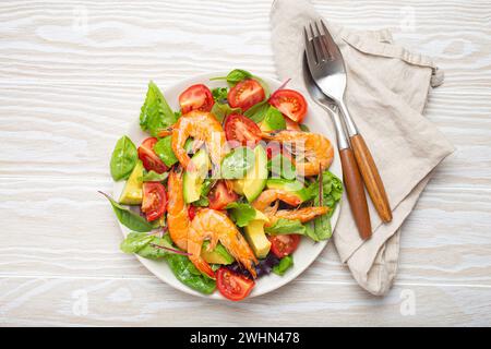 Gesunder Salat mit gegrillten Garnelen, Avocado, Kirschtomaten und grünen Blättern auf weißem Teller mit Besteck auf weißem Holz rustikal Stockfoto